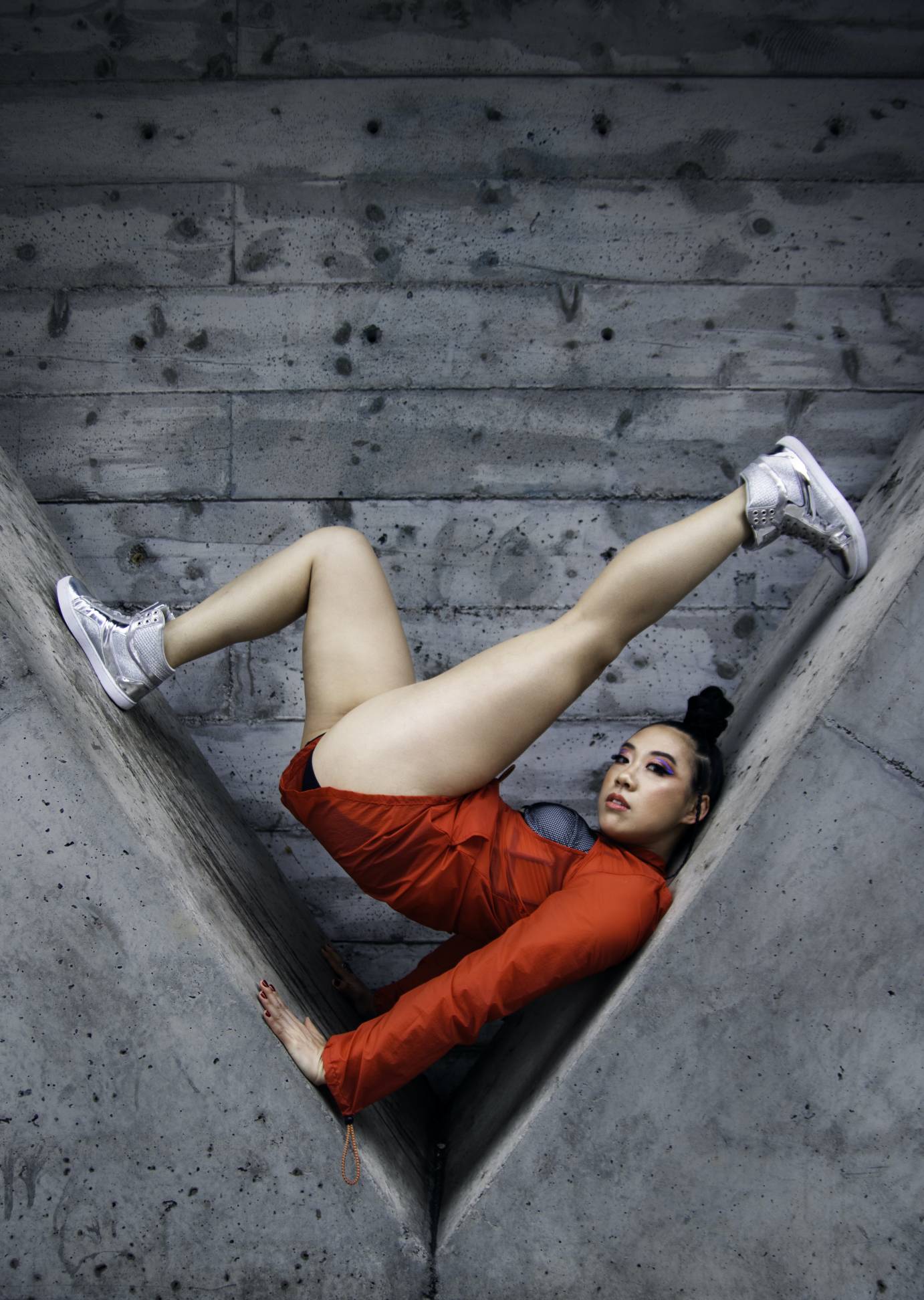 Chinese woman wearing her hair in a bun on top of her head, looking out to the audience. Her body is folded into a cement structure shaped in a V. She wears a long-sleeved red costume. Her legs are bare. She wears above ankle sneakers.